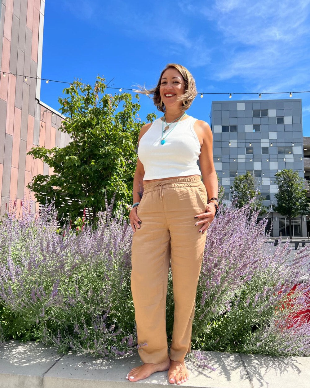 Erika standing barefoot on a low wall in front of tall lavender, hands on her hips, smiling, on a bright summer day.
