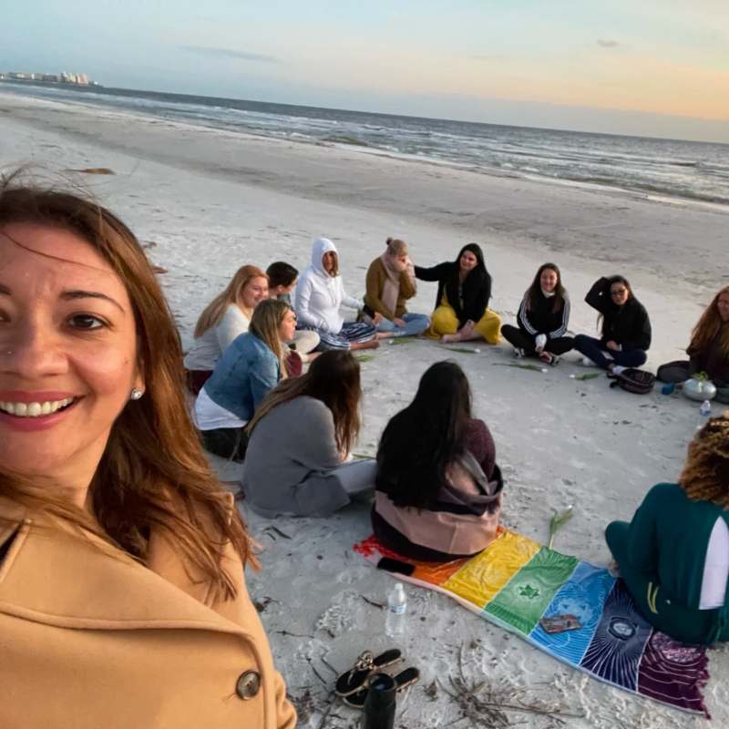 Erika in the foreground smiling, with a circle of women sitting together on the sand at sunset behind her.
