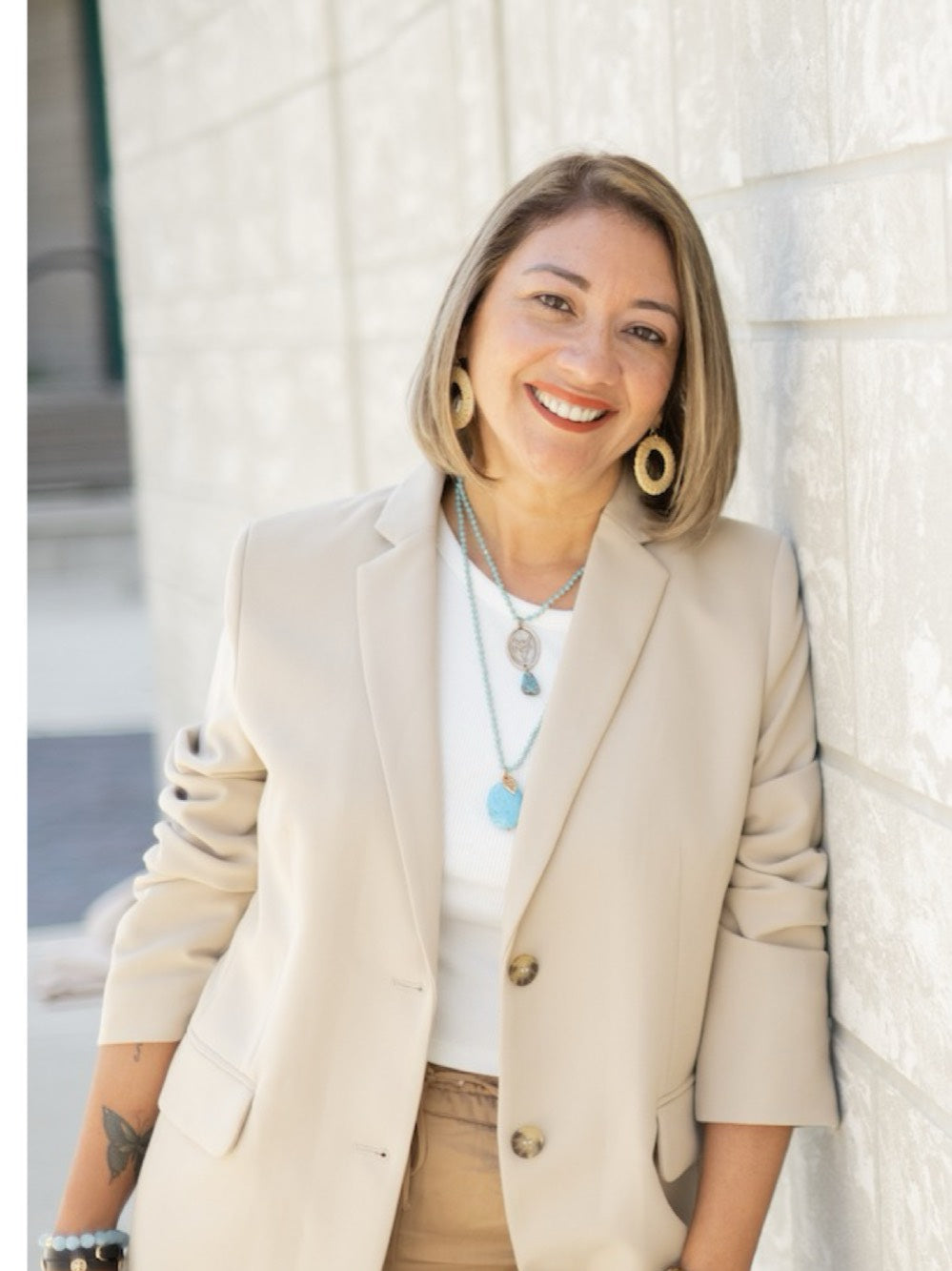 Erika leaning against a white brick wall in a cream blazer, smiling at the camera.
